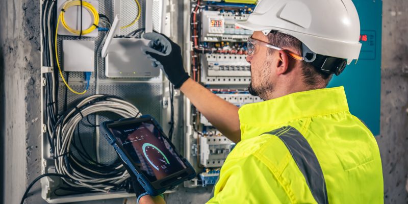 Man, an electrical technician working in a switchboard with fuses. Installation and connection of electrical equipment. Professional uses a tablet.